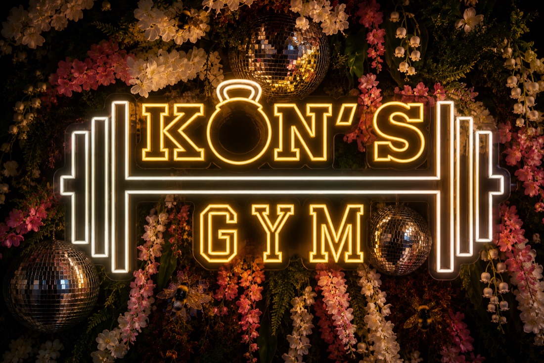 Visualisierung: das Leuchtschild von KON's Gym an einer Blumenwand mit Discokugeln